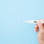 Junge Frau hält weißes digitales Thermometer auf blauem Hintergrund zur Messung der Basaltemperatur schwanger werden Methode