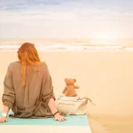 unge alleinstehende Frau mit Teddybär am Strand blickt nachdenklich aufs Meer und überlegt Familiengründung ohne Partner